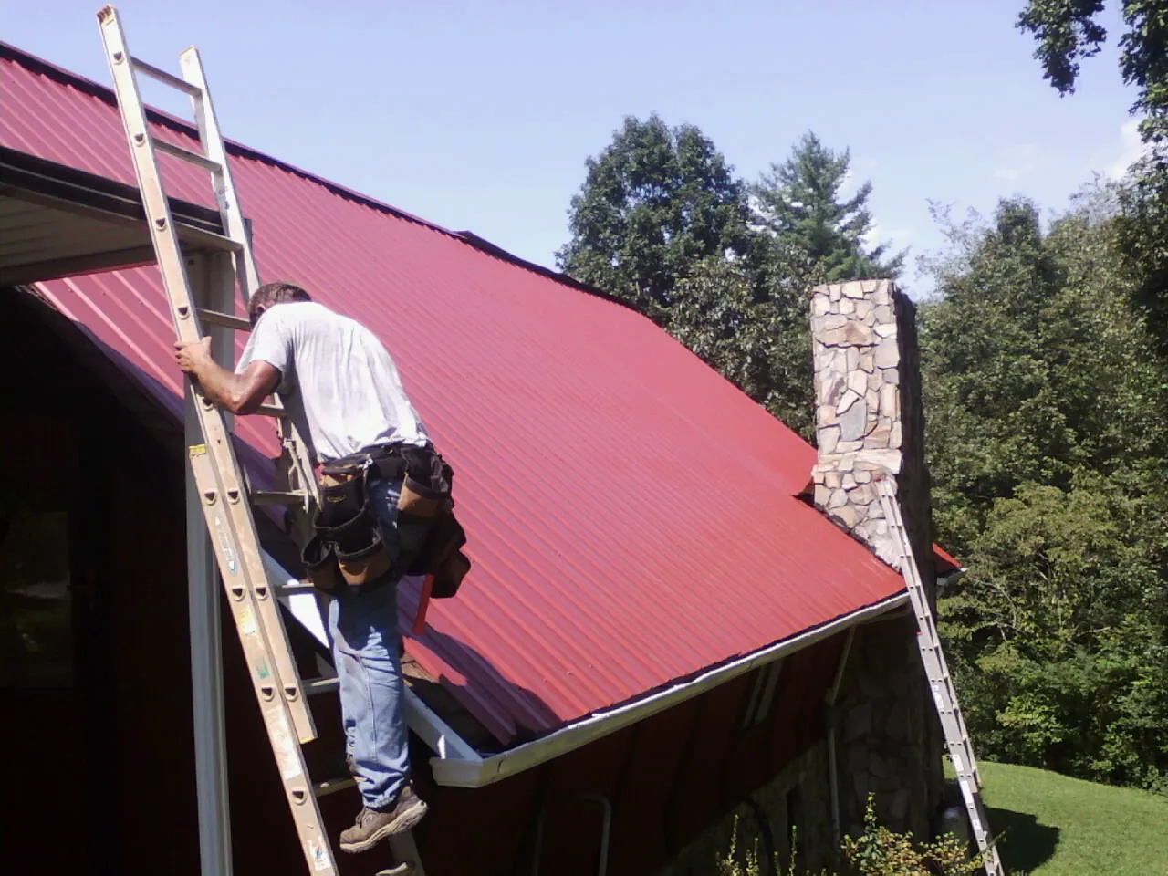 Roofer on a ladder beside a red metal roof and stone chimney — Soffit & Fascia Repair by Roofing Brothers in Crestline